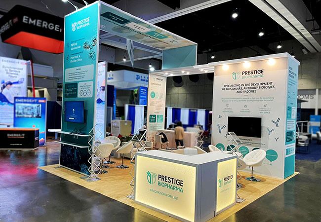 Prestige Biopharma completed booth: an overhead frame above screens, meeting chairs and an angular reception counter