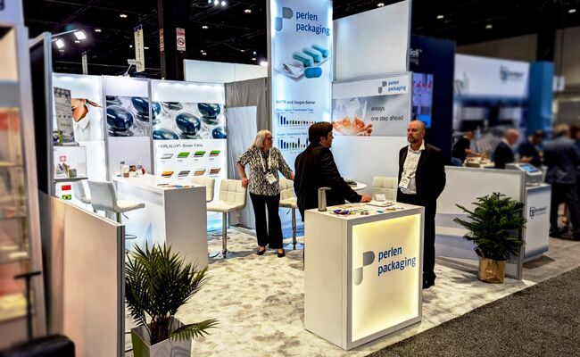 Perlen Packaging completed booth: a hanging sign above product-display panels, a counter and meeting chairs